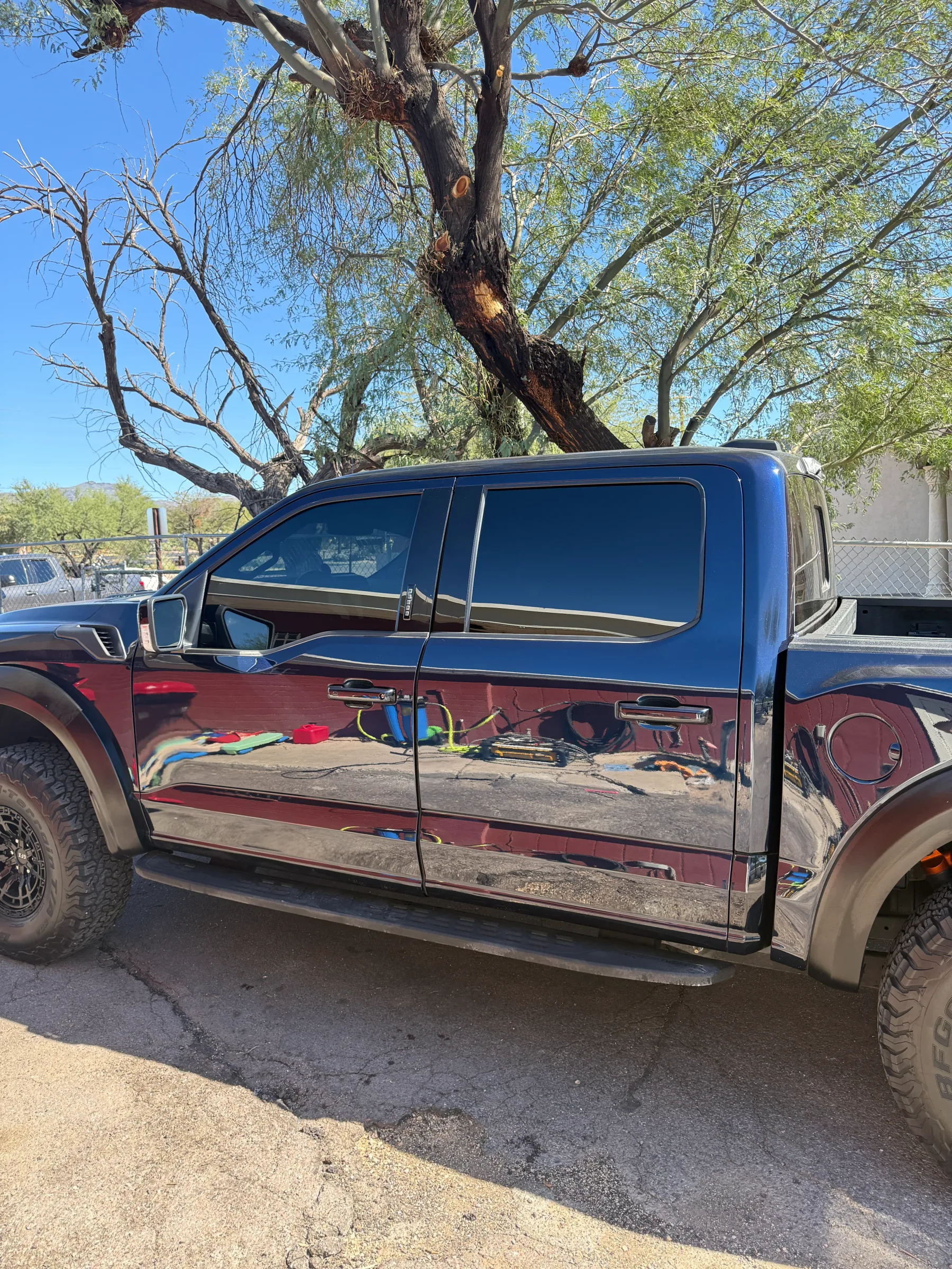 A blue and red pickup truck with tinted windows parked under a large desert tree, showing the contrast between the tinted upper cabin and painted lower body.