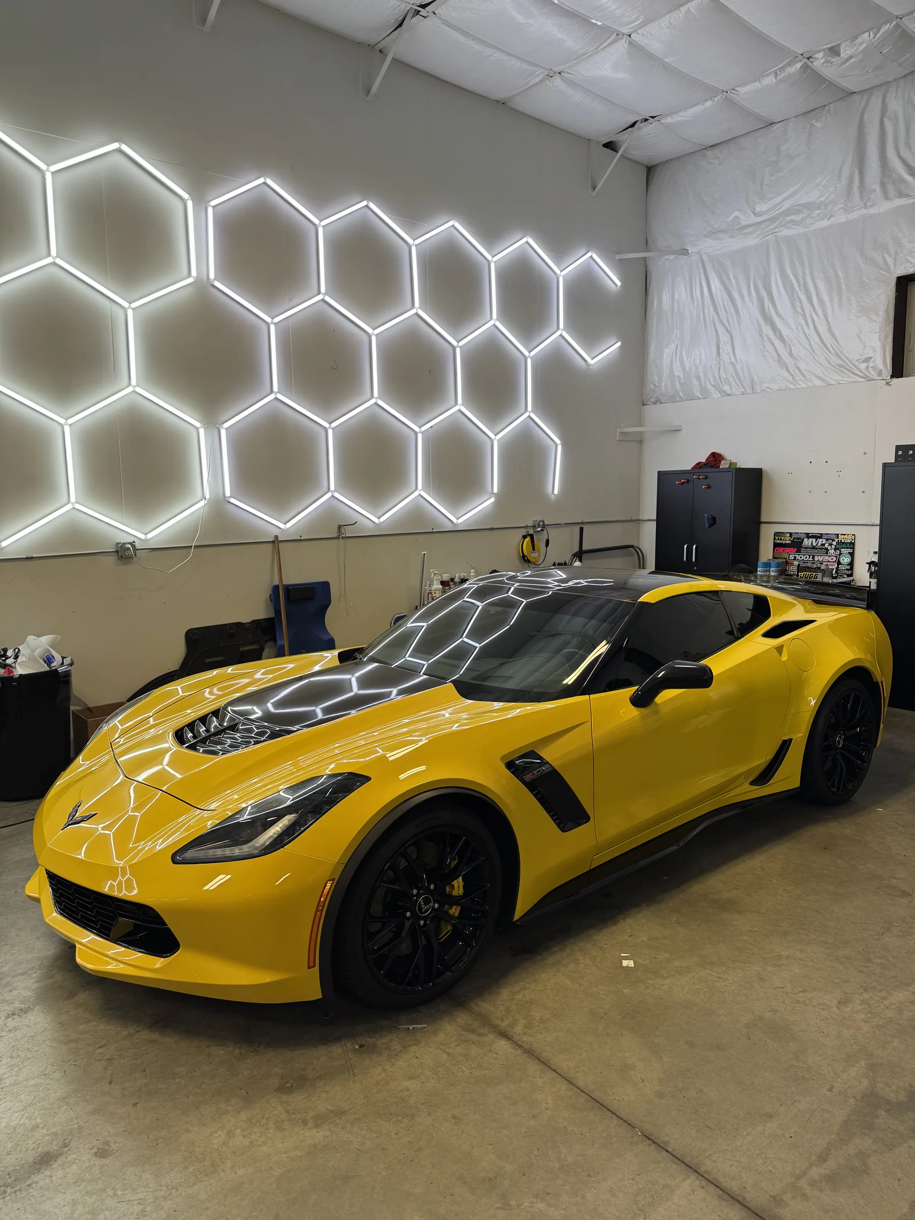 A bright yellow Corvette sports car with white geometric hood wrap and clear paint protection film applied, displayed in a shop with a neon hexagon wall sign.
