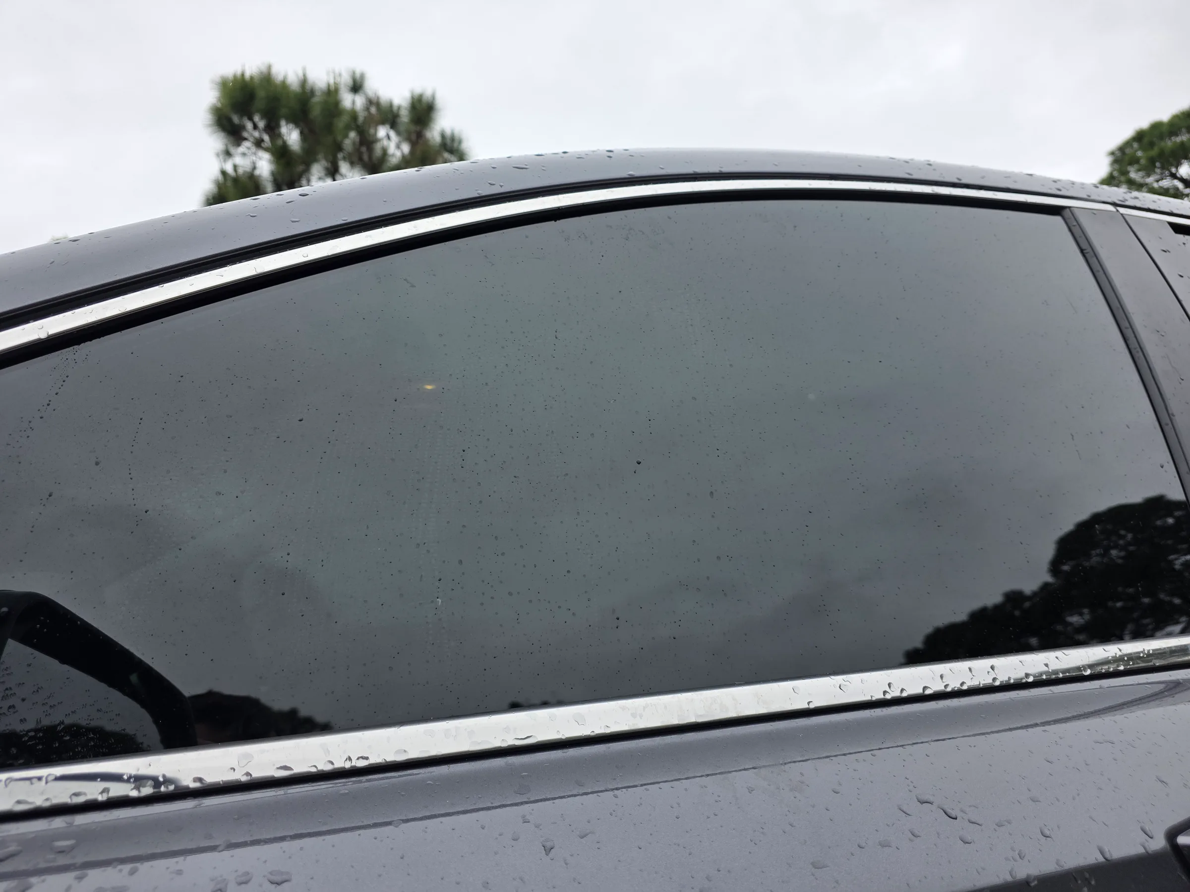 Close-up of a vehicle's rear side window with dark tinted film applied, showing the tint's color and light reflection on a wet surface.