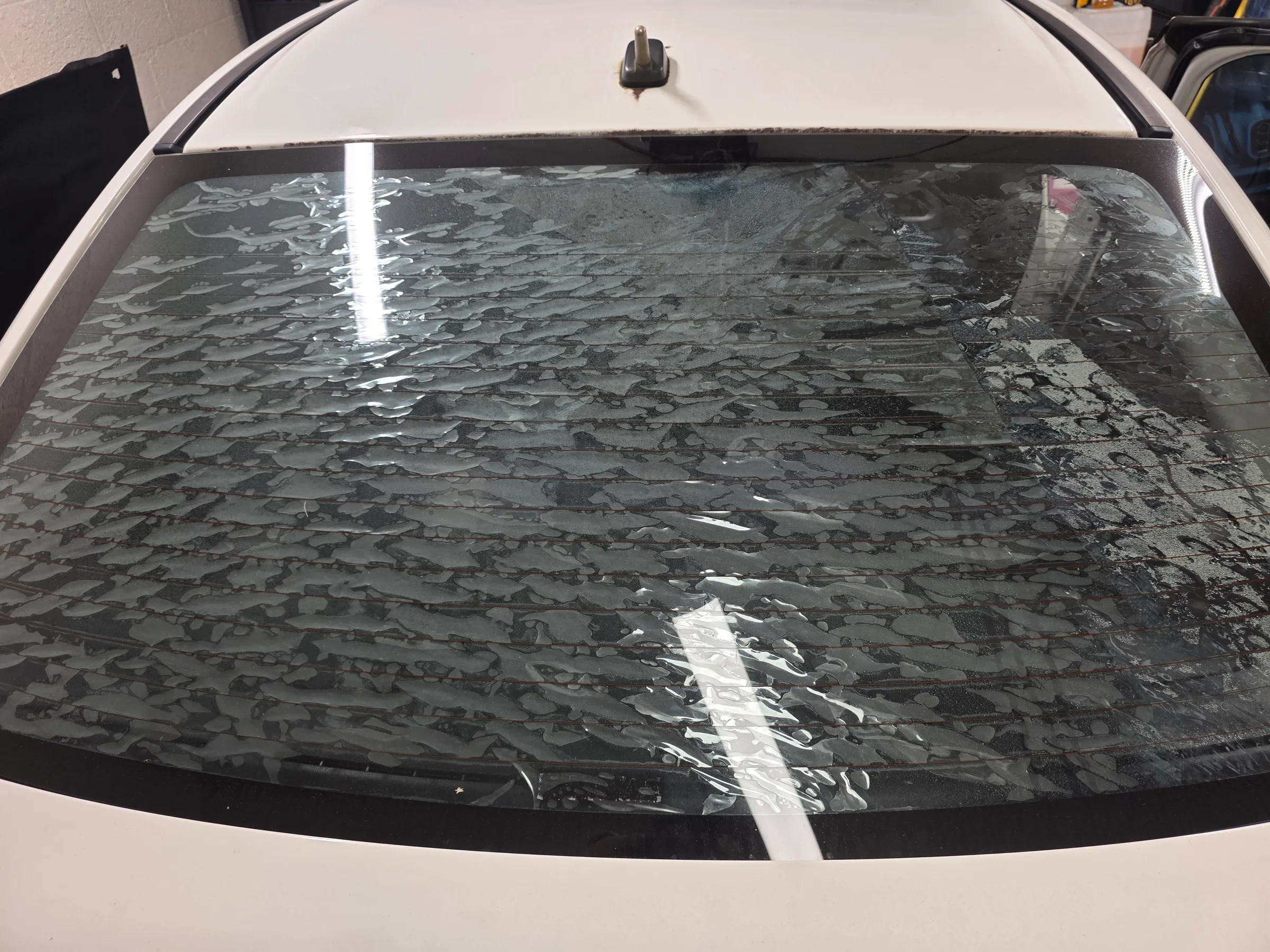A white vehicle's rear windshield with dark window tint applied, showing installation tape and protective film during the application process.