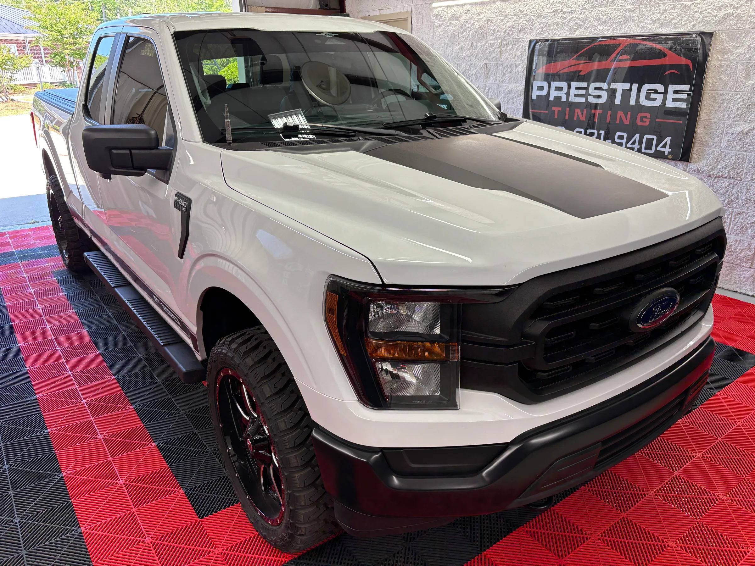 White Ford F-150 pickup truck with dark window tint applied, displayed in a showroom with the Prestige Tinting logo visible on the wall.