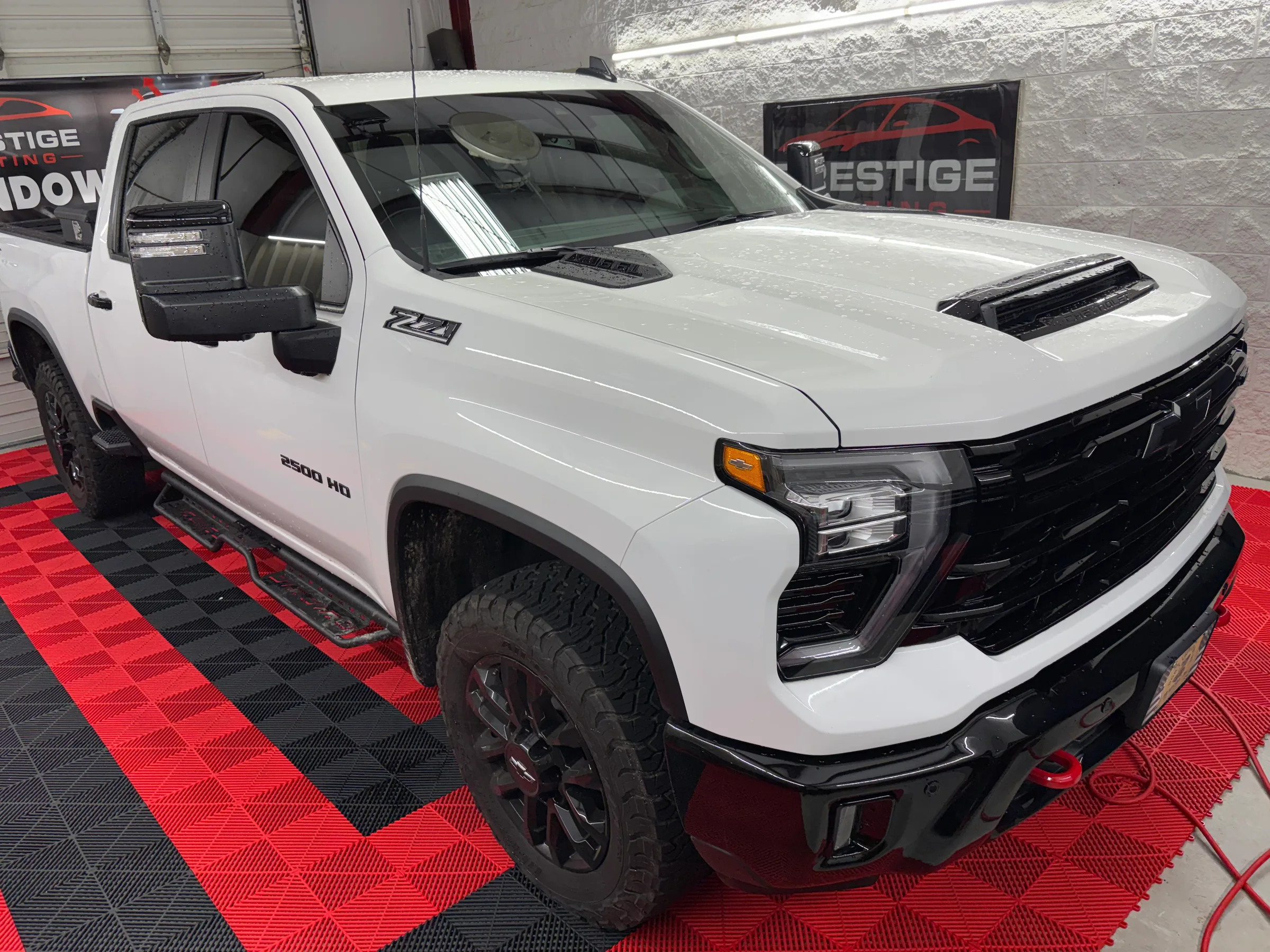 A white Chevrolet Silverado 2500 HD pickup truck with dark window tinting displayed in a showroom with a Prestige Tinting sign visible.