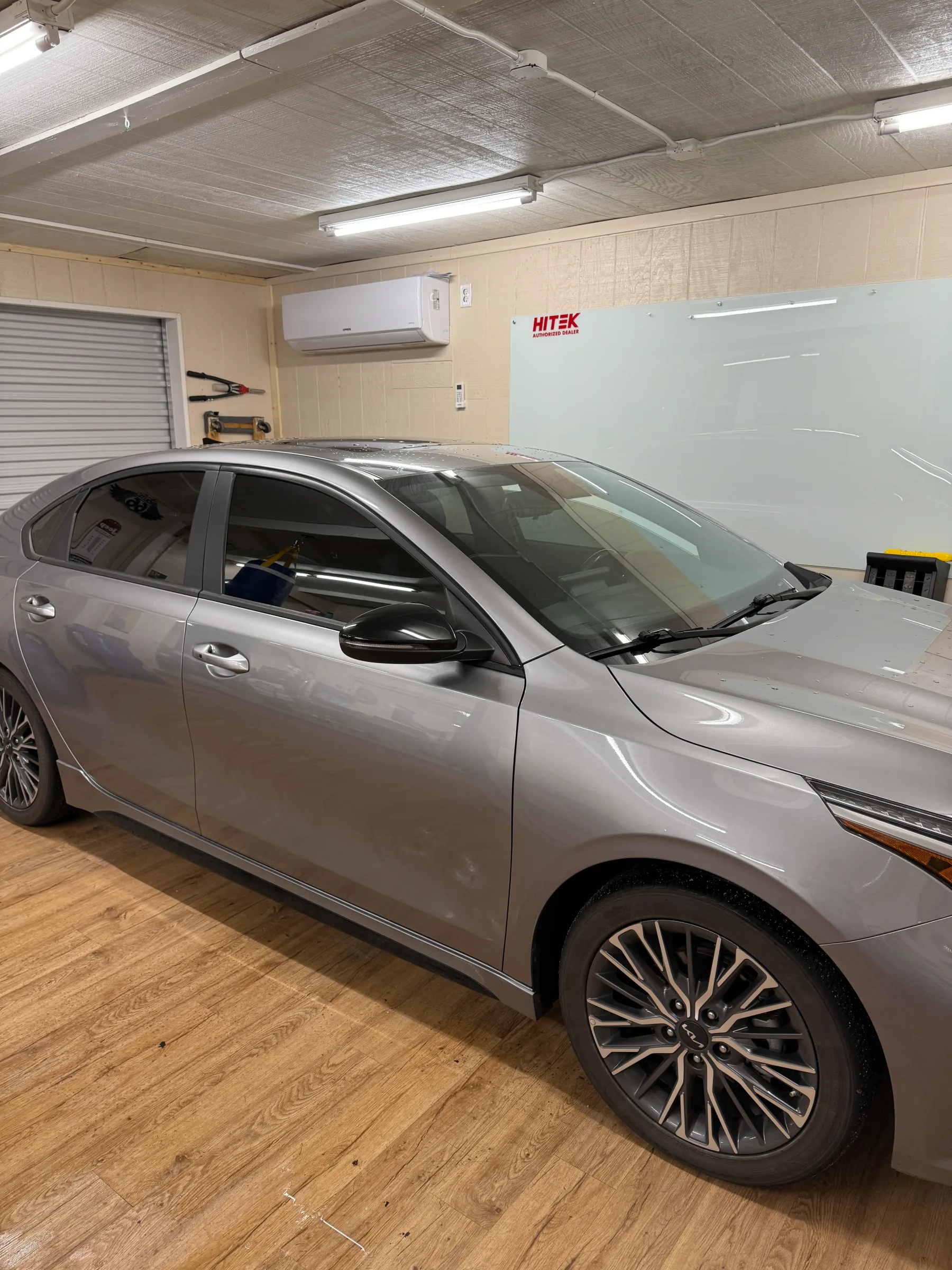 A silver Lexus sedan with freshly applied window tint parked in a professional auto shop with fluorescent lighting and a white tint reference board visible on the wall.