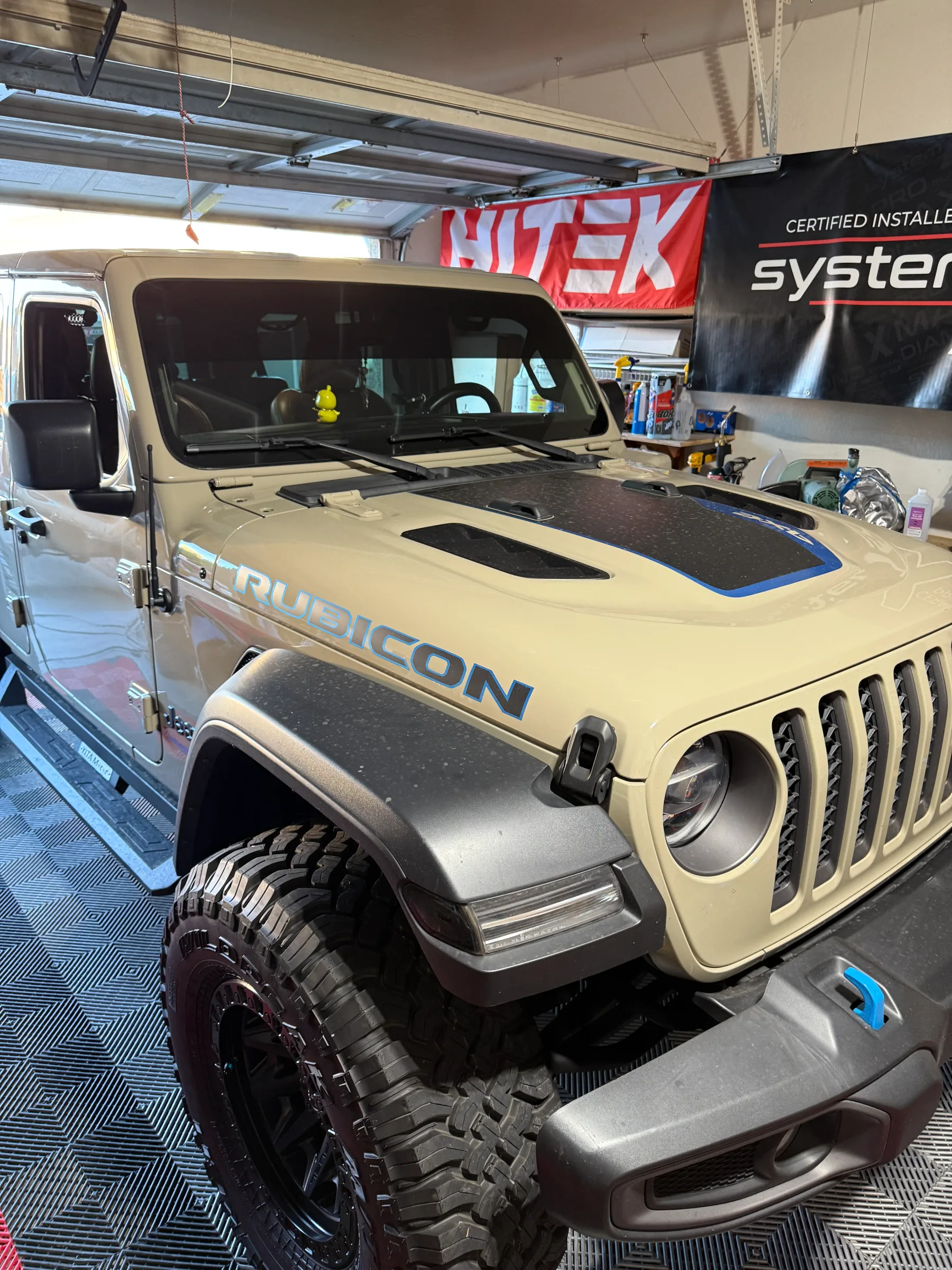 A tan Jeep Rubicon with a two-tone black and blue hood wrap being installed in a professional shop, showing the custom vinyl application on the vehicle's hood.