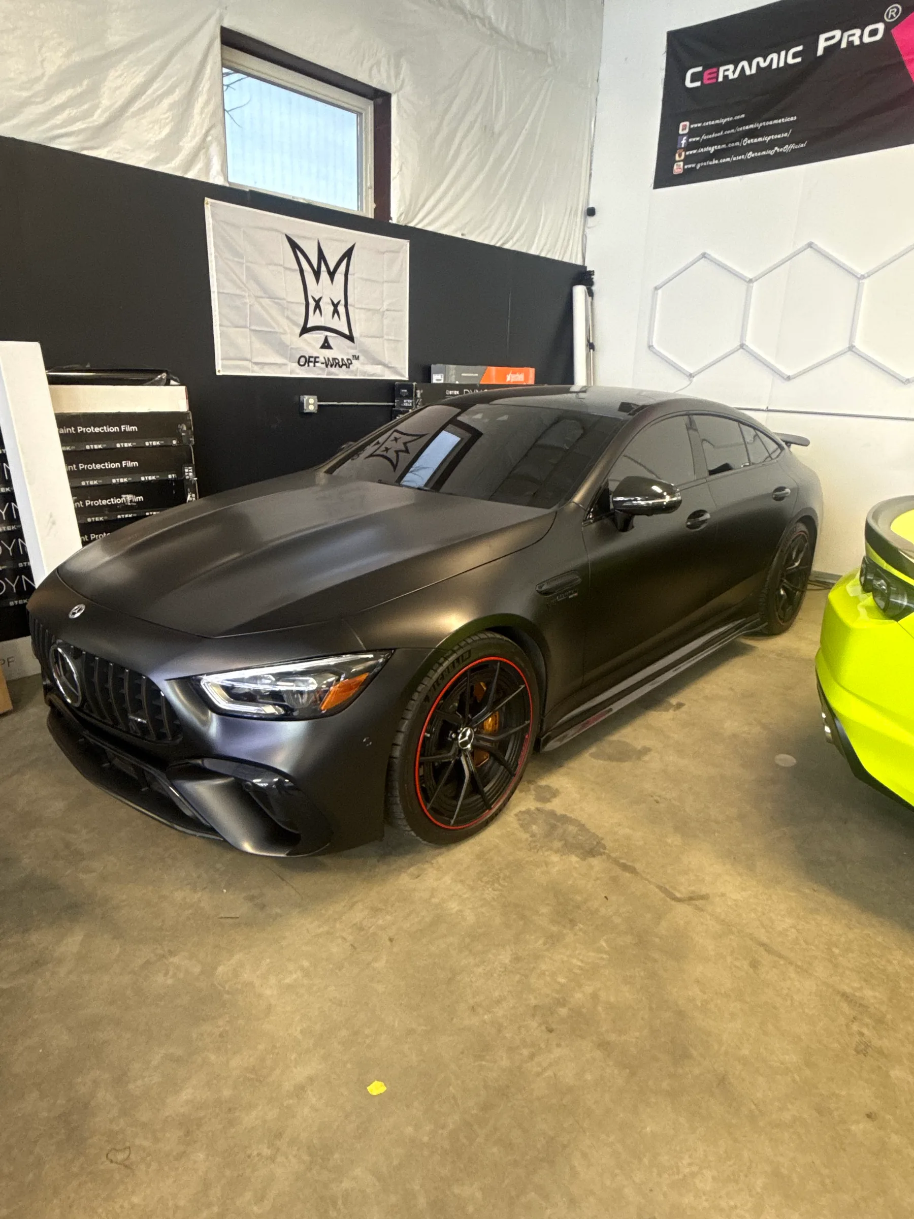 A matte black Mercedes-AMG GT sedan with red accents on the wheels displayed in a shop bay with product branding and banners on the walls.